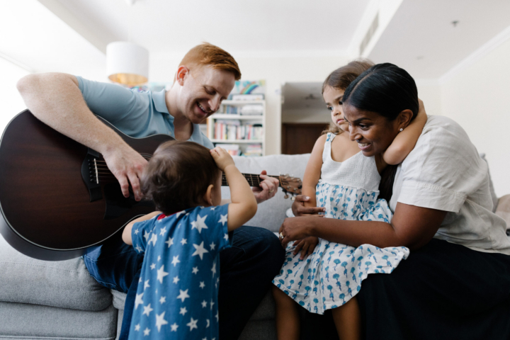 Mother and father smiling with two young girls, father is playing guitar